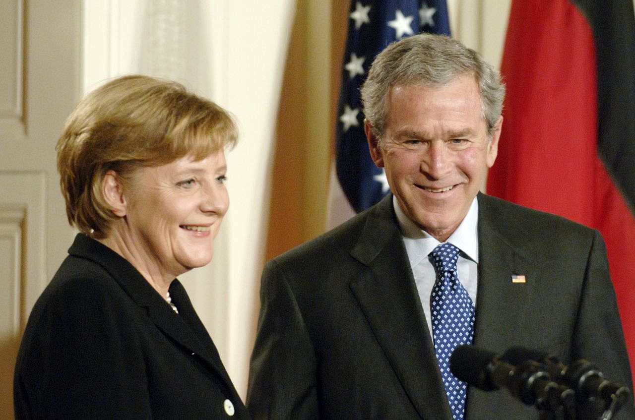 Farbfoto von Angela Merkel und George W. Bush vom Schulterbereich an. Sie stehen im 90-Grad-Winkel zueinander vor Mikrofonen. Im Hintergrund sind Teile der amerikanischen und deutschen Flagge zu sehen.