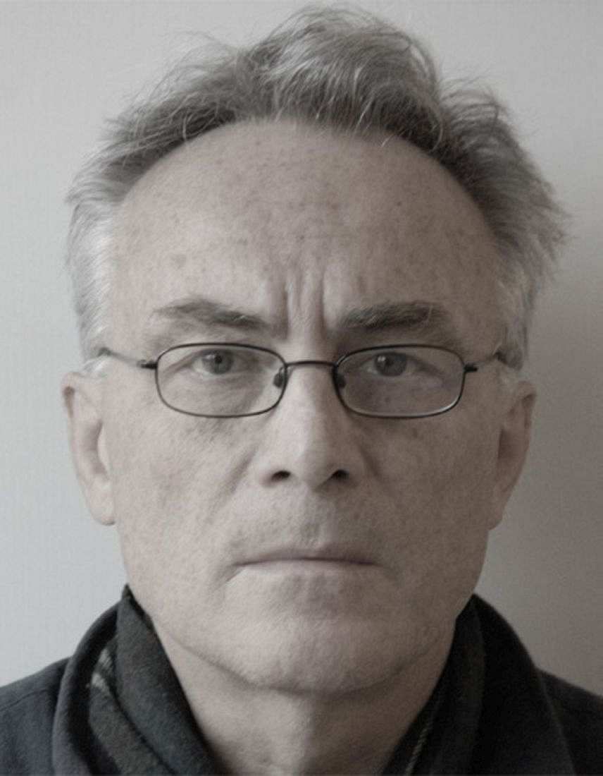 Fotografie. Porträtbild von Norbert Prusko, ca. 70 Jahre alt: graues Haar, schmale schwarze Brille, kein Bart. Prusko trägt einen dunklen Pullover. Im Hintergrund ist eine weiße Wand zu sehen.