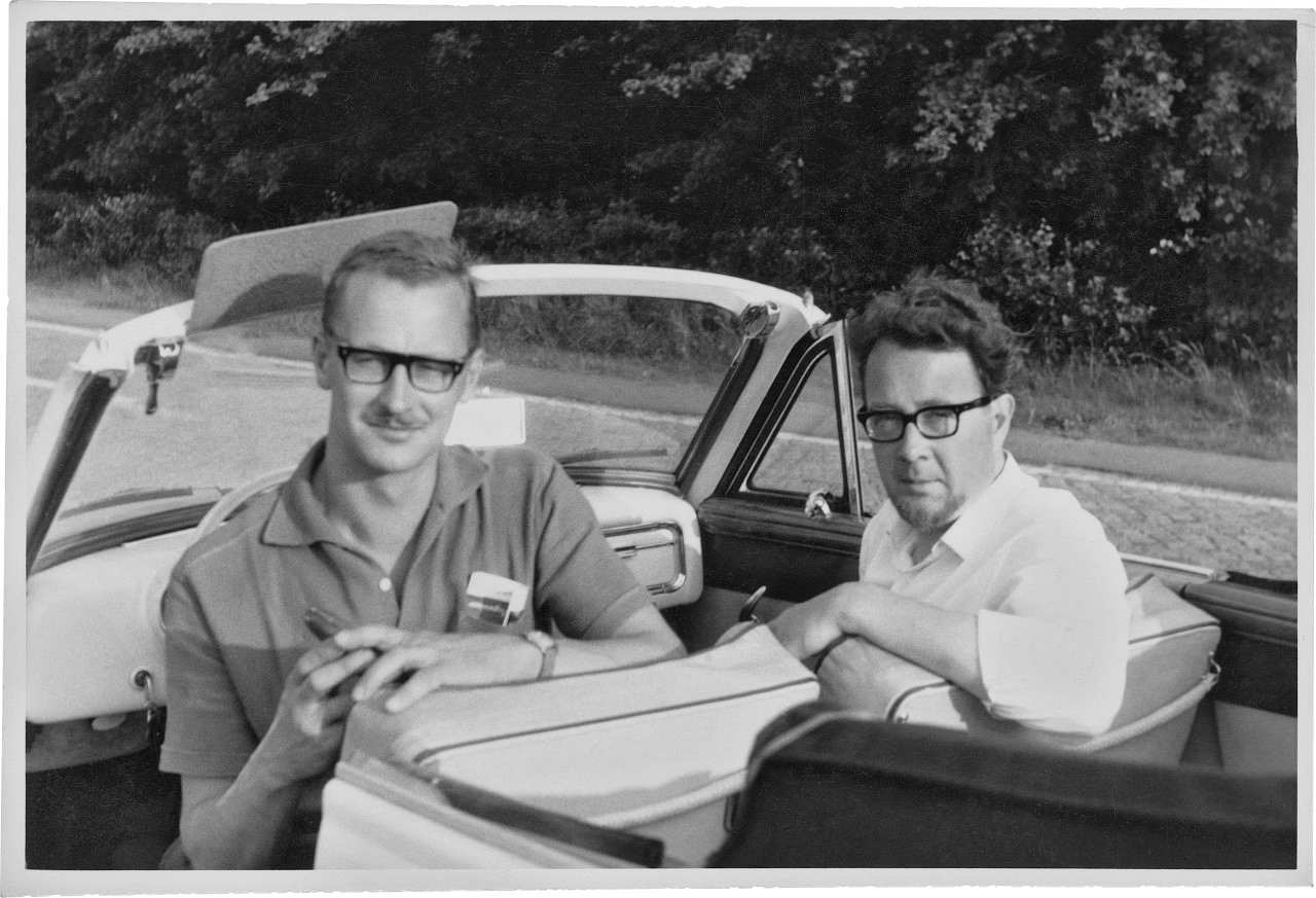 Zwei Männer sitzen in einem Cabriolet und schauen in die hinter ihnen platzierte Kamera. Der linke Mann trägt ein dunkles Poloshirt, eine dunkle Brille und hat einen Schnurrbart. Der rechte Mann trägt ein weißes, hochgekrempeltes Hemd, eine schwarze Brille.