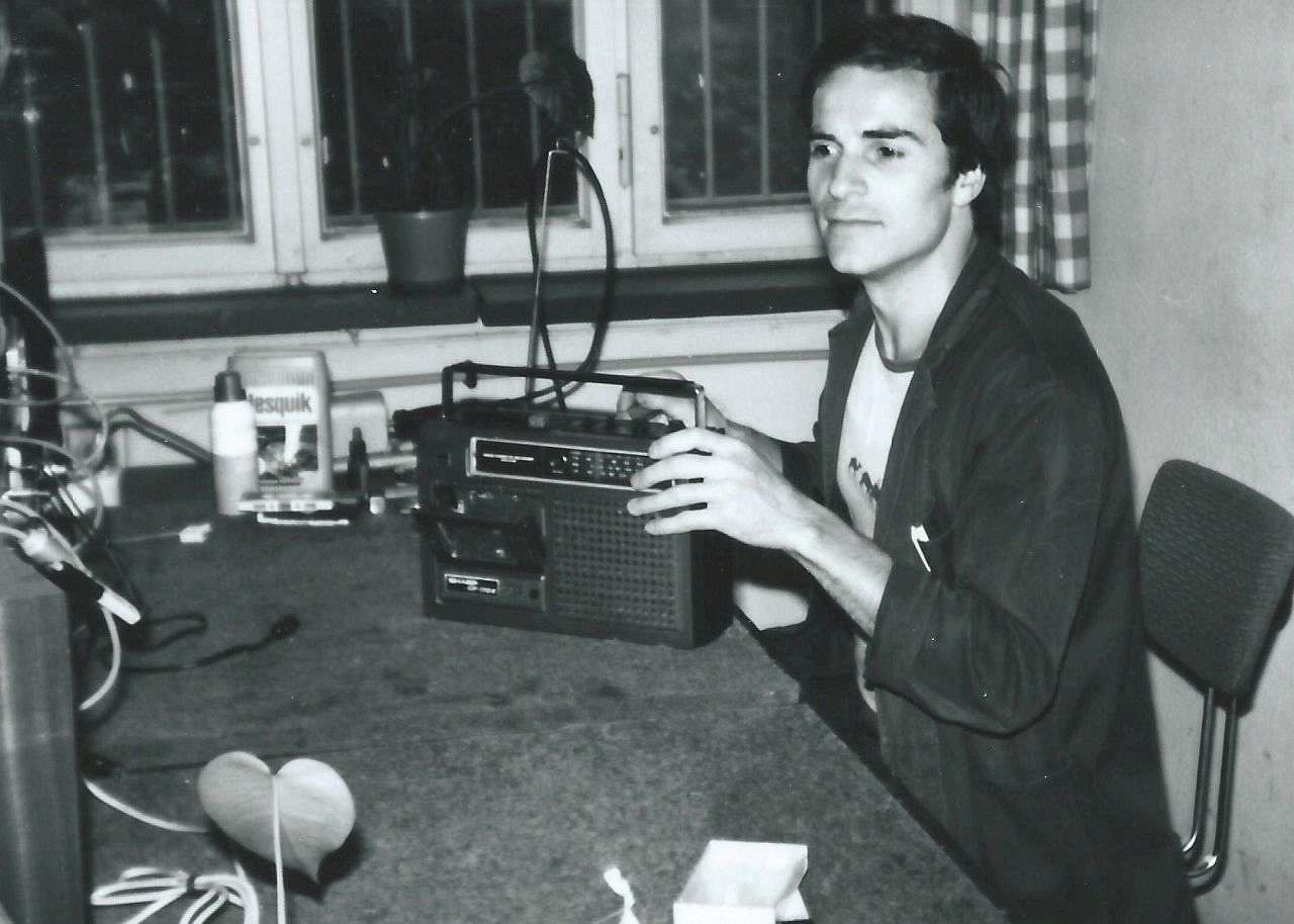 Fotografie, schwarzweiß. Ein junger Mann sitzt an einem Tisch. Vor ihm steht ein Radio. In seiner rechten Hand hält er ein Werkzeug, mit dem er an dem Radio arbeitet. An der rechten Bildseite sieht man die Rückseite von Geräten in denen viele Kabel stecken.