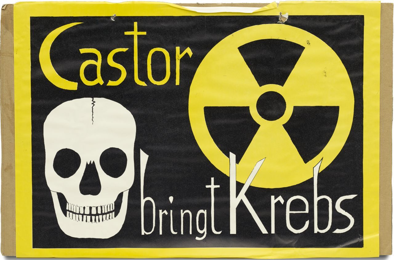 Auf brauner Pappe aufgeklebtes schwarzgrundiges Blatt mit gelber Umrandung; unten links Totenschädel, oben rechts Symbol für Radioaktivität; Text oben links 'Castor' und unten rechts 'bringt Krebs'. Am Rand oben zwei Löcher mit einer durchgezogenen Kordel.