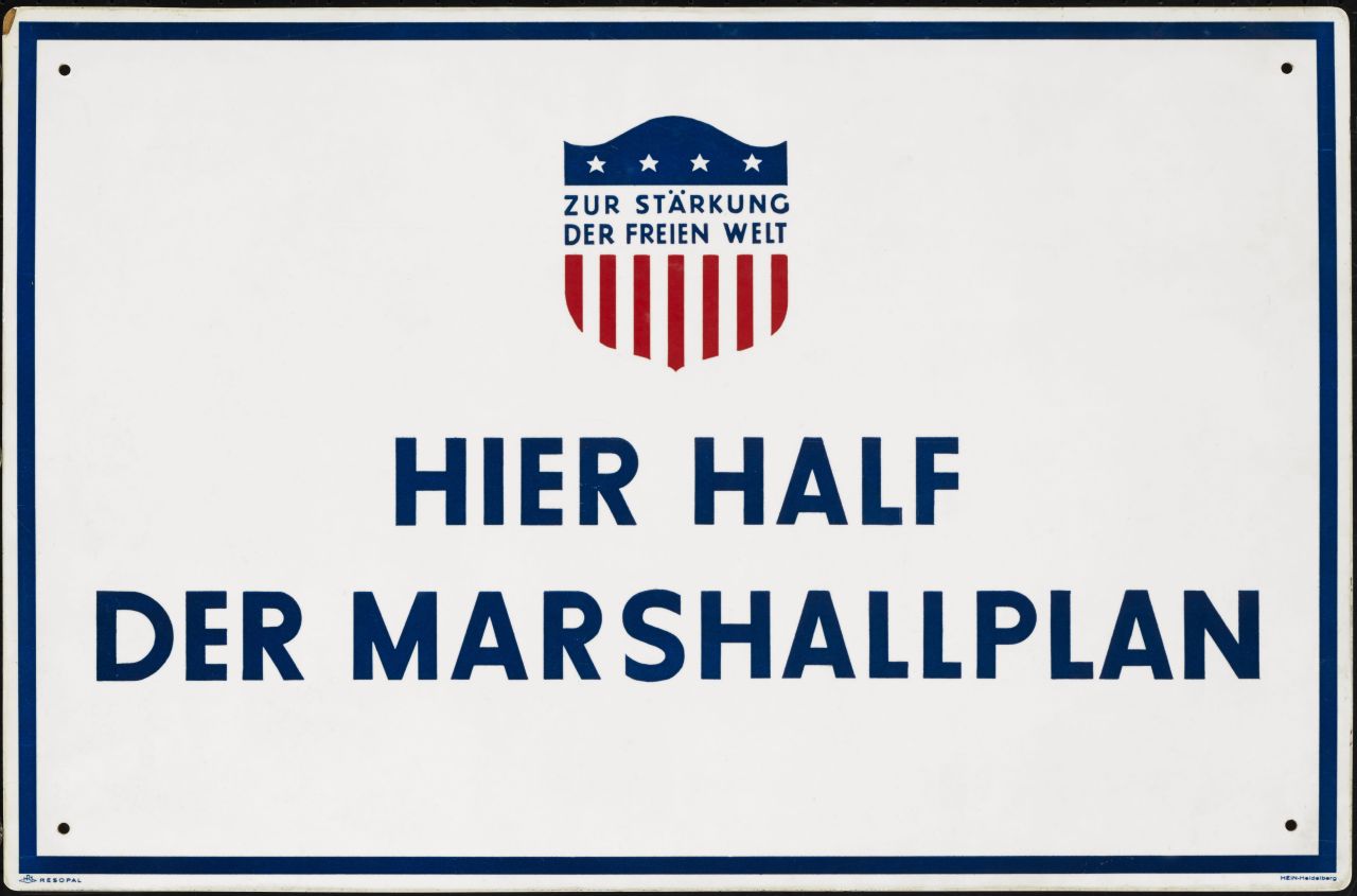 Weißes Emailleschild mit blauer Umrandung, oben Mitte Wappen mit Stars and Stripes mit Text: Zur Stärkung der freien Welt, blau, rot, weiß, Text: blaue Großbuchstaben, darunter: Hier half der Marshallplan. In den Ecken je ein Loch zur Befestigung.