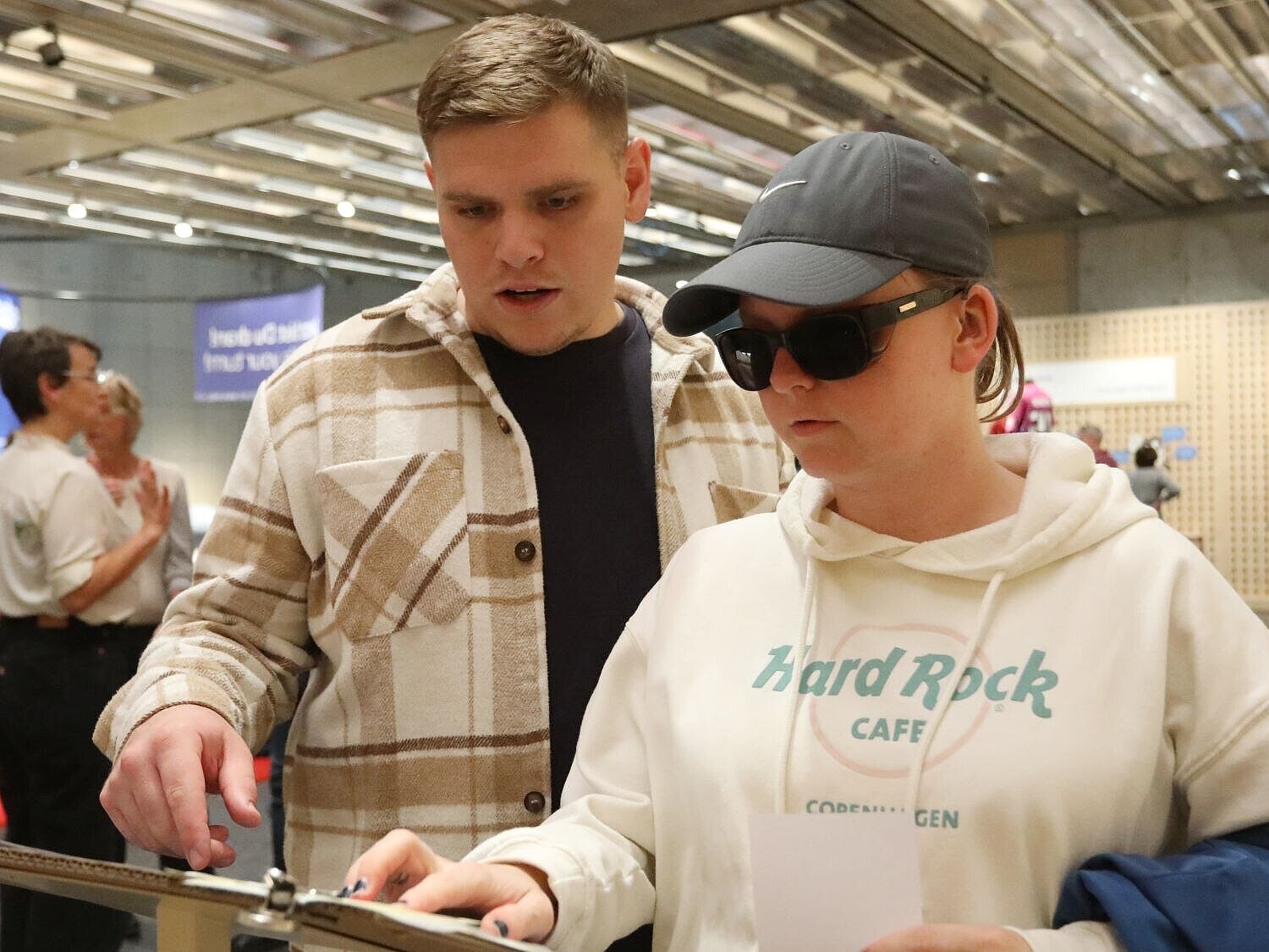 Eine junge Frau mit Sonnenbrille legt ihre Hand auf eine Tafel. Neben ihr steht ein junger Mann.