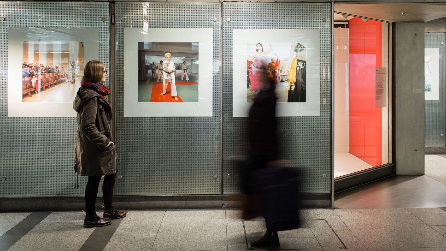 In einer U-Bahn-Station steht eine junge Frau vor einer Wand mit drei großformatigen Fotos und betrachtet sie, eine weitere Frau läuft mit einem Koffer durch das Bild 
