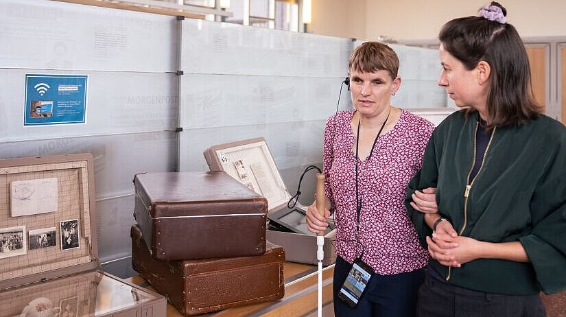 Eine Besucherin mit Blindenstock hört den AudioGuide und geht mit einer Begleiterin durch die Ausstellung