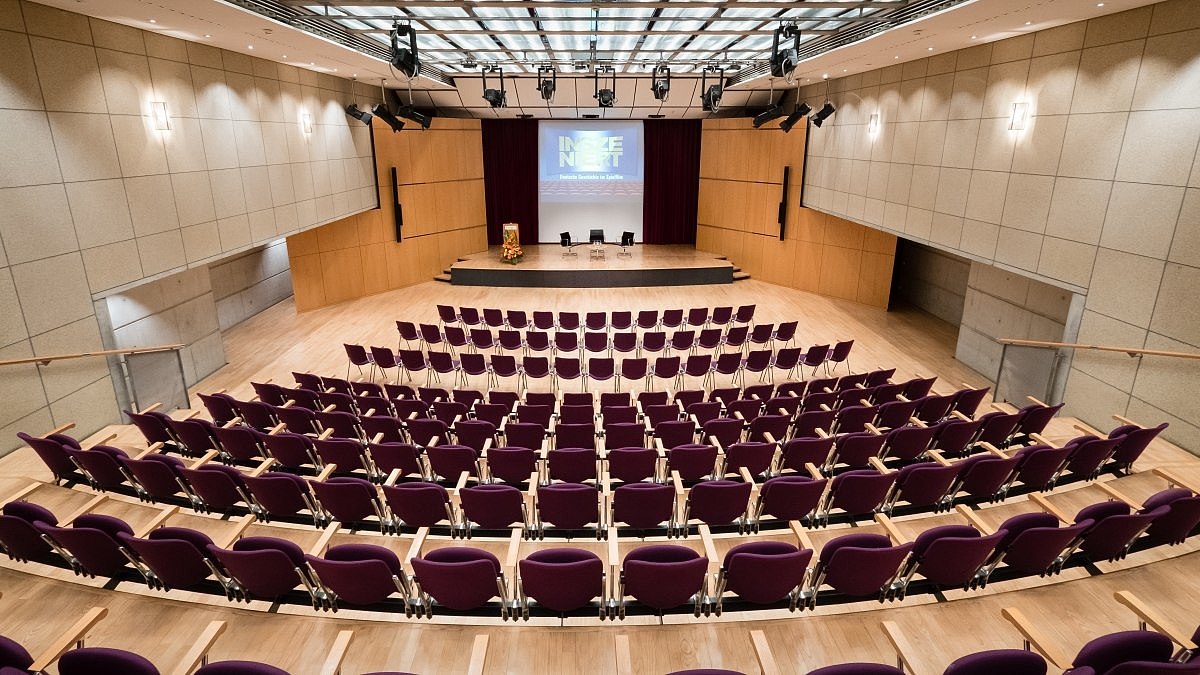 Blick in einen großen Raum mit Stuhlreihen und einer Bühne mit Podium.