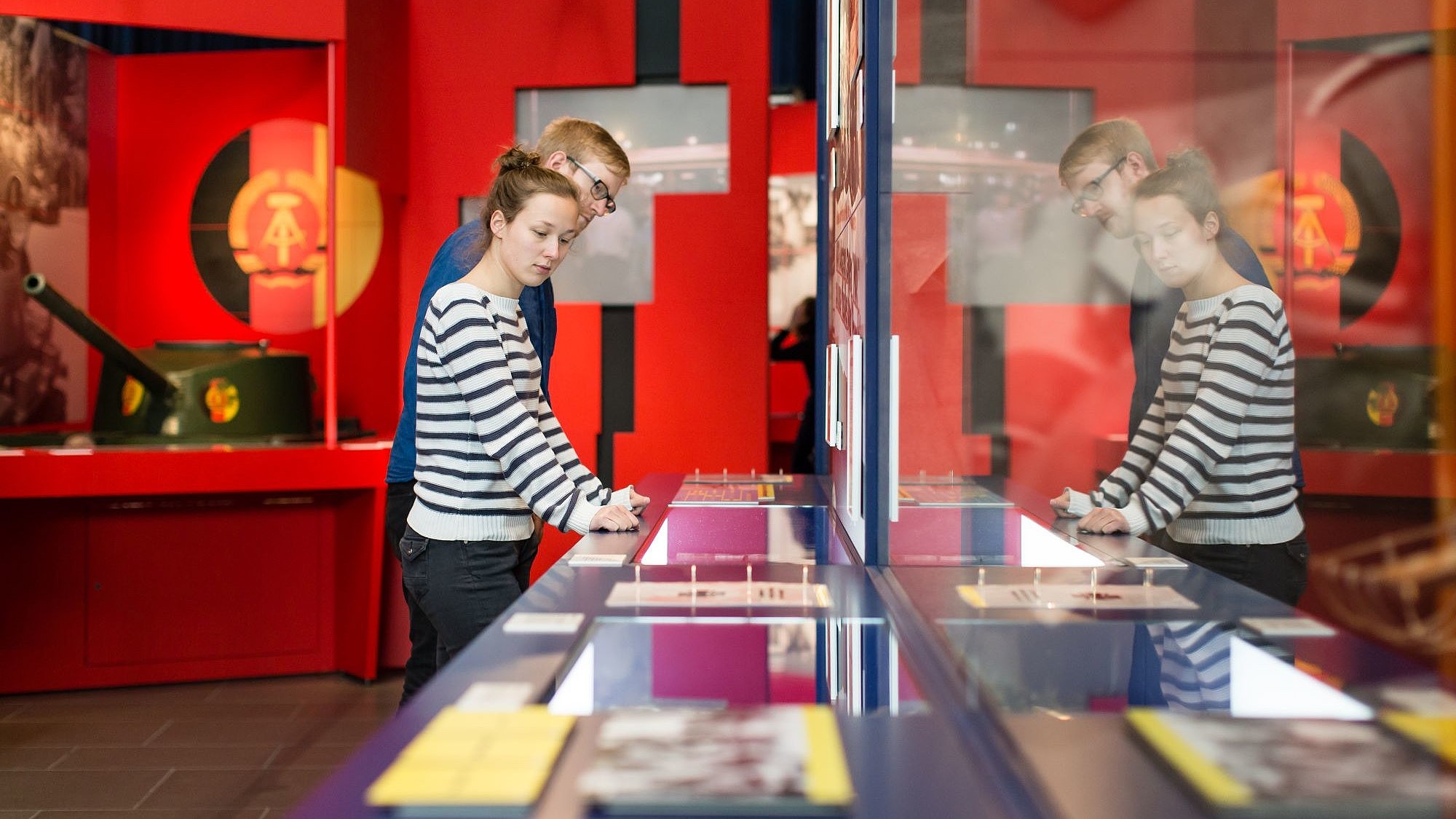 Blick in einen Ausstellungsraum mit einer großen Längsvitrine im Vordergrund und roter Ausstellungsarchitektur im Hintergrund mit DDR- Wappen und einem Kinder-Panzer. Eine junge Frau und ein junger Mann sehen sich die Objekte in der Vitrine an