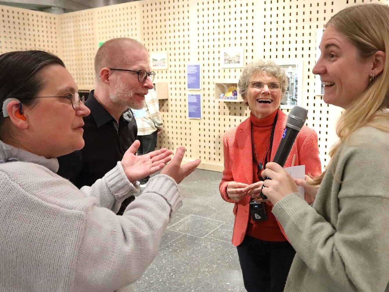 Eine Frau mit Mikrofon steht mit zwei anderen Frauen und einem Mann in einem kleinen Kreis. Die drei sind ihr zugewandt. Eine Frau spricht mit ihr und hebt gestikulierend die Hände, die andere lacht sie fröhlich an.