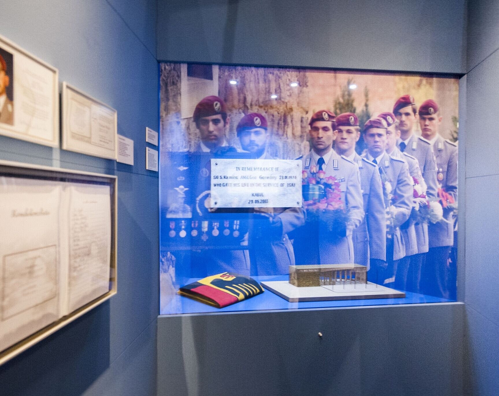 Blick in eine Ausstellungseinheit mit einem großformatigen Foto von Soldaten in einem Trauerzug, einem Architekturmodell, einer zusammengefalteten Deutschlandflagge, einer Traueranzeige und weiteren Dokumenten