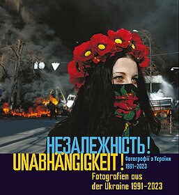Ausstellungsplakat mit ukrainischer und deutscher Schrift, im Hintergrund ist ein Foto einer Frau mit Blumenkranz im Haar und Halstuch mit Blumenaufdruck zu sehen. Hinter ihr sieht man brennende Barrikaden und Autos. 