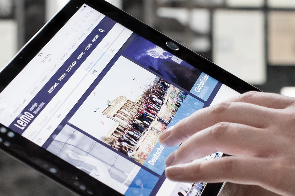 Auf einem Tablet wird eine Website angezeigt, man kann ein Foto mit Menschen auf der Berliner Mauer vor dem Brandenburger Tor erkennen. Eine Hand bedient das Tablet.