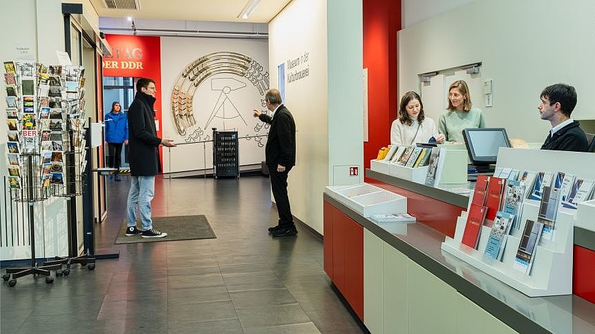 Besucherinnen und Besucher mit Aufsichtspersonal am Infotresen und im Eingangsbereich der Ausstellung. 