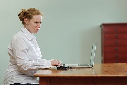 Blinde Frau sitzt am Tisch mit Laptop und angeschlossener Braillezeile, Foto: dzb lesen
