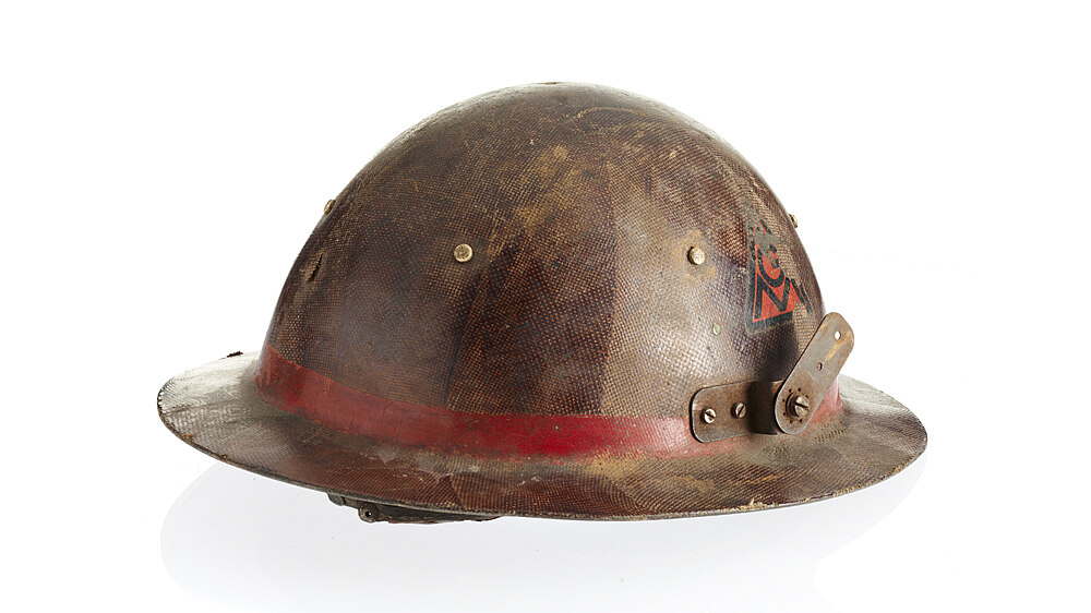 Braun-roter Helm mit flacher, schmaler Hutkrempe. Außen am Übergang von Krempe zu Helmaufsatz roter Streifen. An beiden Seiten angeschraubte Metallverstrebungen, vermutlich zur Befestigung weiterer Schutzvorrichtungen.