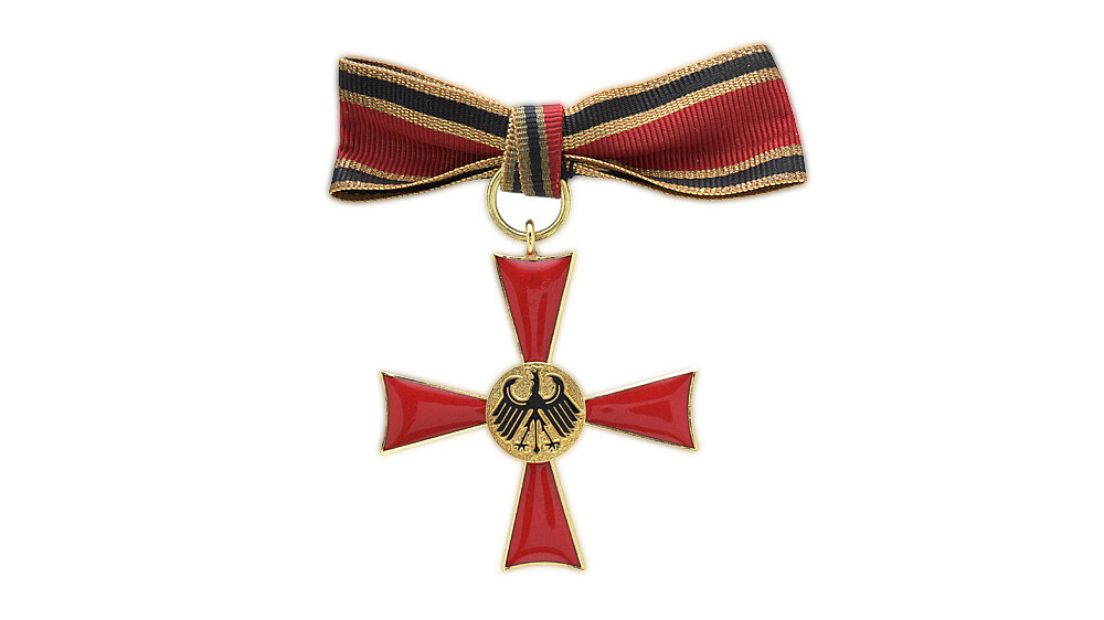 Goldfarben eingefaßtes, rot emailliertes Kreuz mit goldfarbenem Medaillon. Darauf schwarzer erhabener Bundesadler. Auf dem oberen Kreuzarm Öse mit Bandring. Rote Damenschleife mit gold-schwarz-goldfarbenen Seitenstreifen.