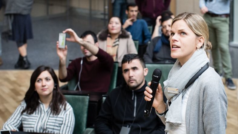 Im Vordergrund eine junge Frau mit Mikrofon, dahinter sitzen junge Erwachsene in der Ausstellung, hören zu und machen Fotos