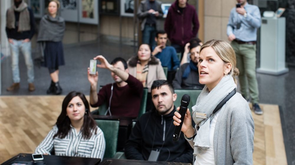 Im Vordergrund eine junge Frau mit Mikrofon, dahinter sitzen junge Erwachsene in der Ausstellung, hören zu und machen Fotos