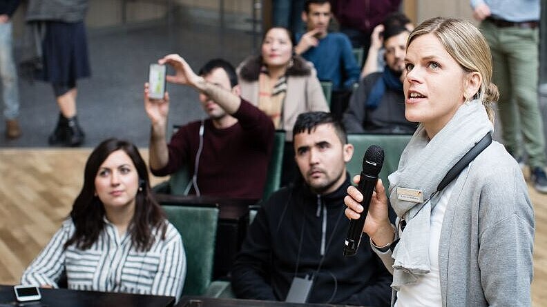 Im Vordergrund eine junge Frau mit Mikrofon, dahinter sitzen junge Erwachsene in der Ausstellung, hören zu und machen Fotos