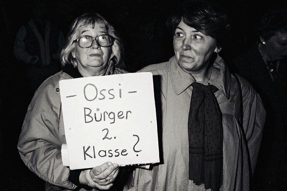 Zwei Frauen, eine davon trägt ein Plakat mit der Aufschrift - Ossi - Bürger 2. Klasse?