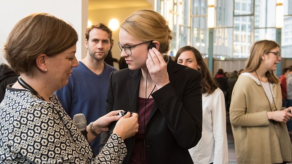 Eine junge Frau befestigt den Ohrstecker den Audio-Gruppenführungssystems, eine andere Frau hilft ihr dabei