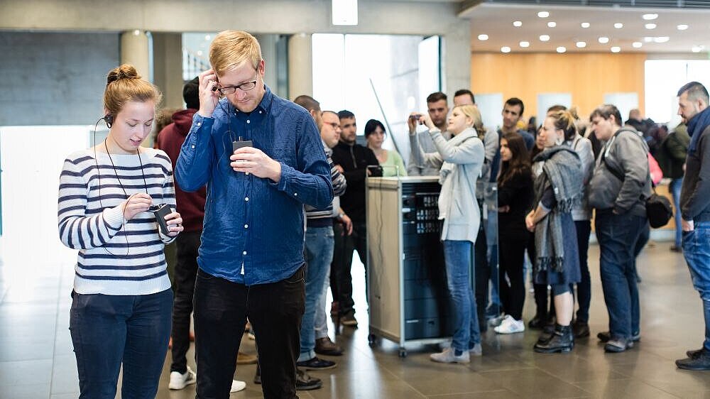 Im Vordergrund eine junge Frau und ein junger Mann, die ihre Geräte und die Ohrhörer für die Begleitung anbringen, im Hintergrund weitere Personen bei der Ausgabe der Geräte