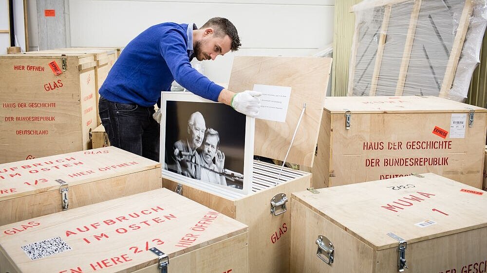 Ein junger Mann verstaut ein großeformatiges, gerahmtes Foto in einer offenen Holzkiste. Um ihn herum stehen weitere geschlossene Holzkisten