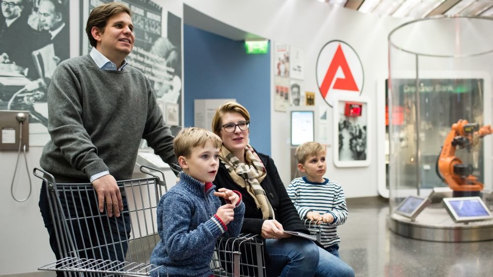 Eine Junge und seine Mutter sitzen in einem Ausstellungsraum auf Stühlen in Form von Einkaufswagen, der Vater steht dahinter, ein anderer kleiner Junge daneben