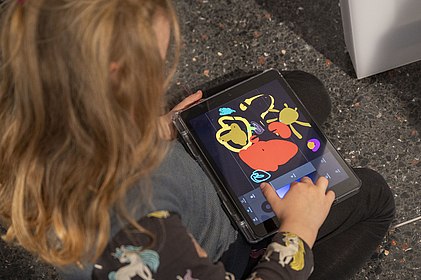 Mädchen mit blonden Haaren malt auf einem Tablet mit bunten Farben