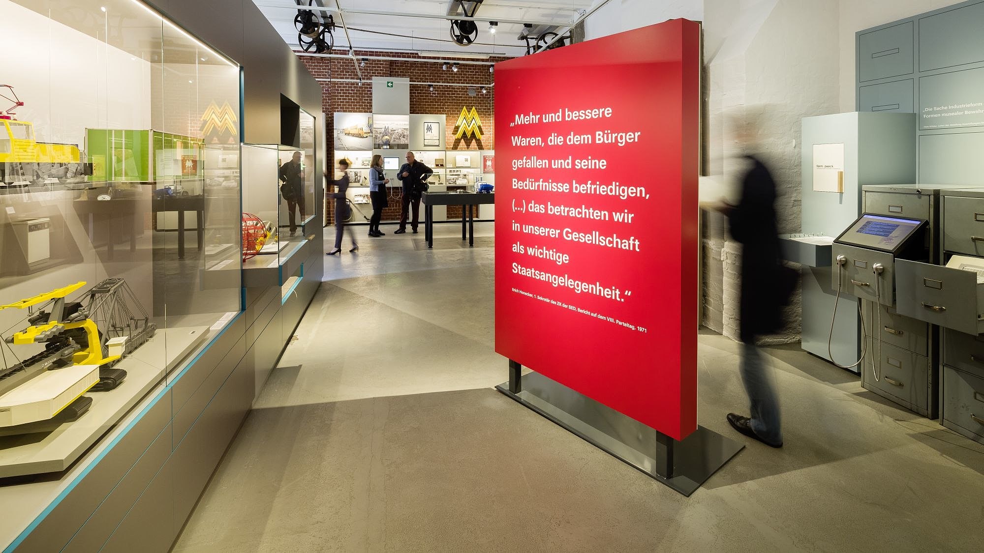 Blick in einen großen Ausstellungsraum, in der Mitte ein großer roter Raumtrenner mit einem Zitat von Erich Honecker, links Modelle von Nutzfahrzeugen in einer Vitrine