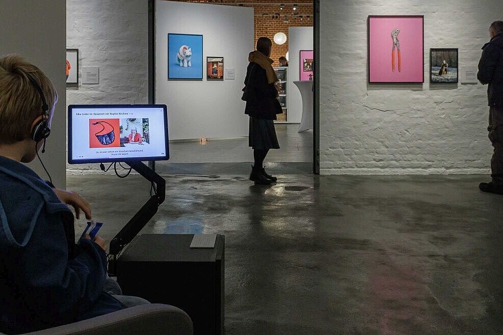 Junge mit Kopfhörern vor einem Monitor. Im Hintergrund befinden sich Fotografien in der Ausstellung. 