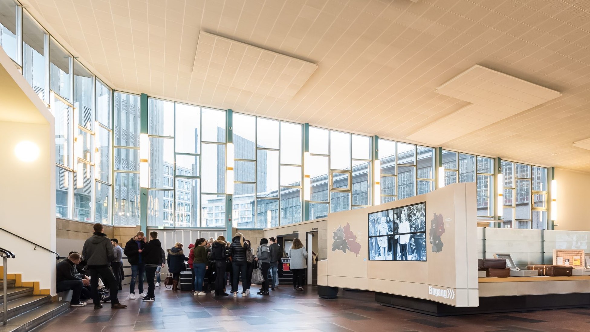 Blick in die große ehemalige Ausreisehalle und den Eingangsbereich der Ausstellung. Besucher stehen vor der großen Fensterfront, hinter der man u.a. den Bahnhof Friedrichstraße sieht