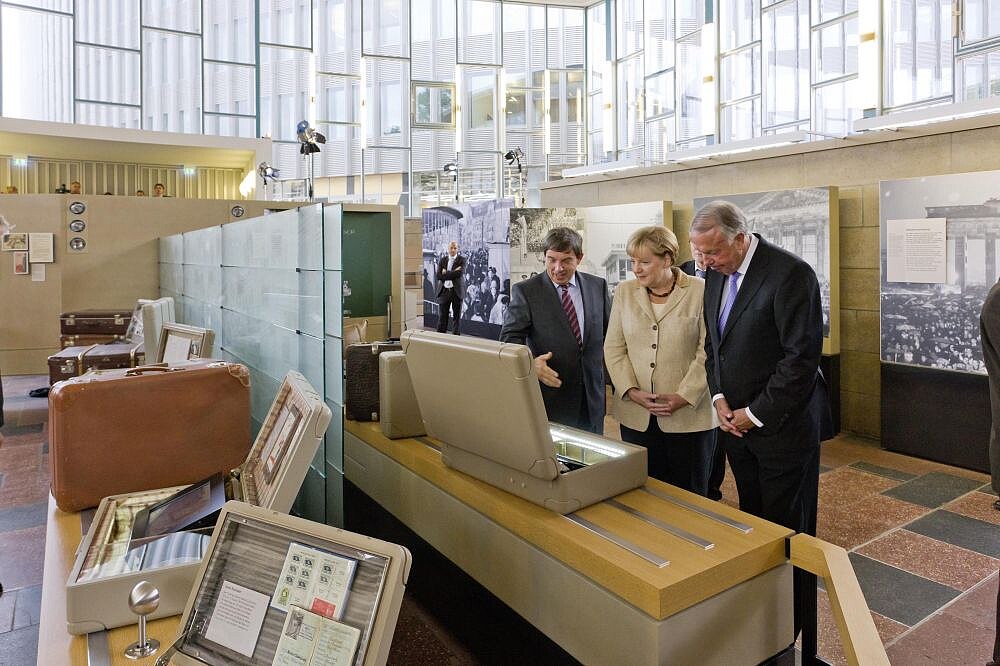 Blick in die Ausstellung des Tränenpalastes, Stiftungspräsident Hütter erläutert Bundeskanzlerin Merkel, Kulturstaatsminister Neumann den Inhalt eines Koffers, im Hintergrund die große Glasfassade des Tränenpalastes