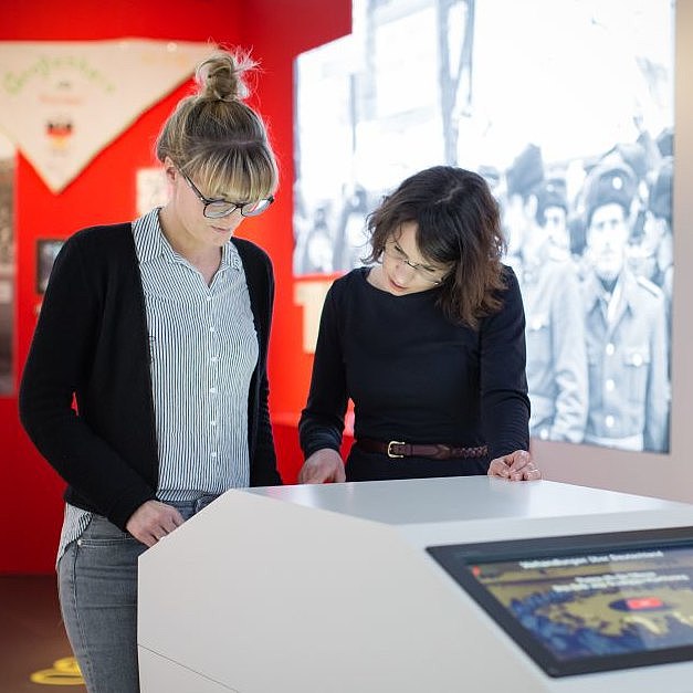 Zwei junge Frauen stehen in einem Ausstellungsraum über einen Monitor gebeugt