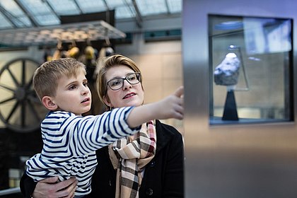 Ein kleiner Junge auf dem Arm seiner Mutter zeigt auf einer Vitrine, in der ein etwa faustgroßer Gesteinsbrocken präsentiert wird