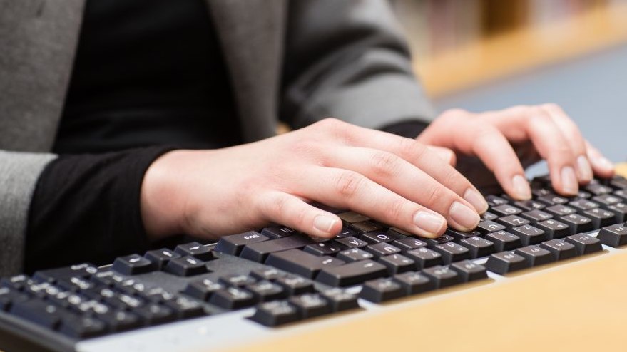 Zu sehen sind der Oberkörper einer Frau und Hände auf einer Computertastatur, im Hintergrund ein Bücherregal