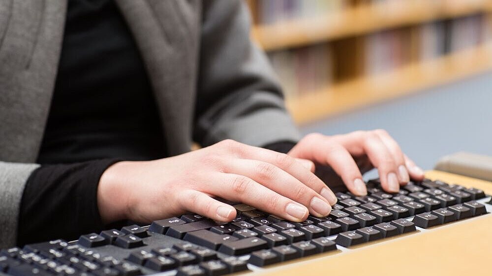 Zu sehen sind der Oberkörper einer Frau und Hände auf einer Computertastatur, im Hintergrund ein Bücherregal