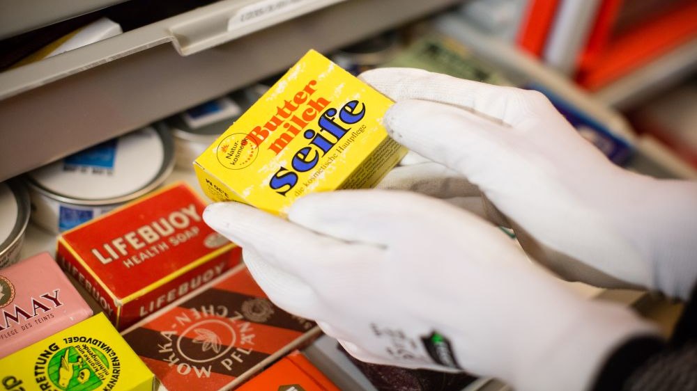 Zwei Hände in weißen Handschuhen halten eine kleine gelbe Pappschachtel mit der Aufschrift Buttermilchseife. Darunter liegen in einer Schublade weitere bunte Schachteln.