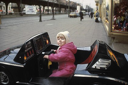 Titelbild zur Ausstellung. Kleines Mädchen sitzt in einem schwarzen Spielauto.