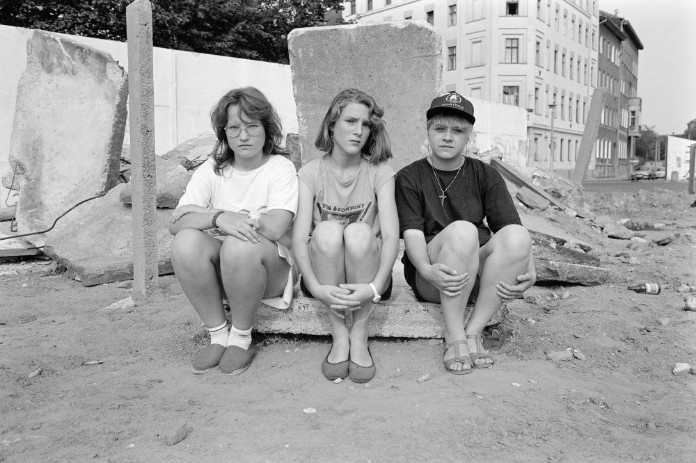 Auf dem Schwarz-weiß Foto sitzen 3 Mädchen vor einem Stück Betonmauer. 