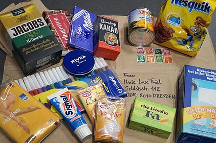 Der Inhalt eines Westpakets Nahrungsmittel, Hygieneprodukte und Filzstifte liegen auf Packpapier mit einer Dresdner Adresse.