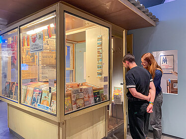 Ein Besucher und eine Besucherin einer Gruppe betrachten Zeitschriften in einem Kiosk in der Ausstellung.