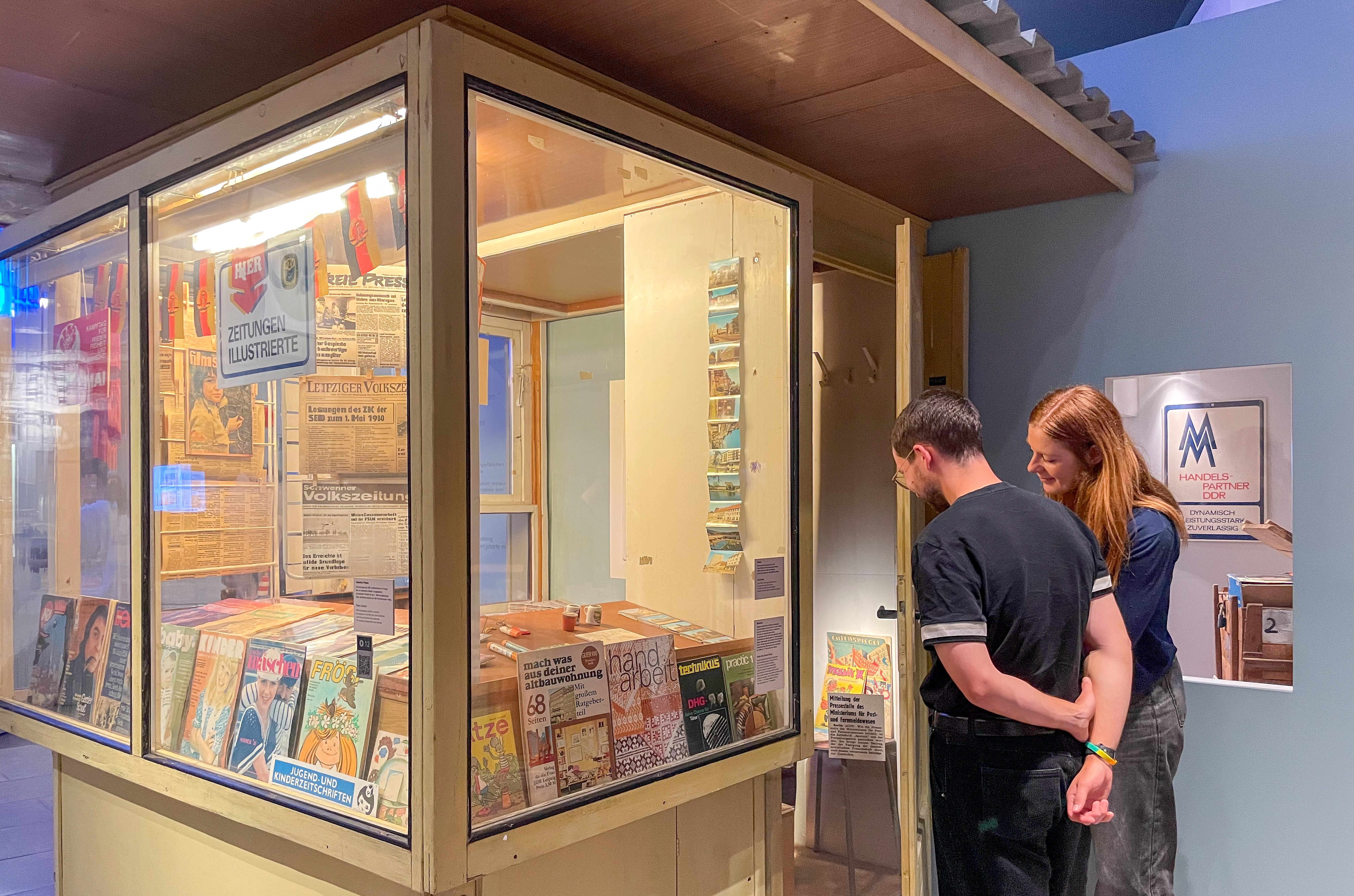Ein Besucher und eine Besucherin einer Gruppe betrachten Zeitschriften in einem Kiosk in der Ausstellung.