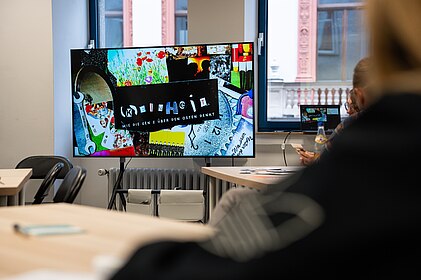 Foto eines Bildschirms in einem Klassenzimmer. Auf dem Bild ist eine Collage mit dem Titel "(k)einheit" zu sehen. 