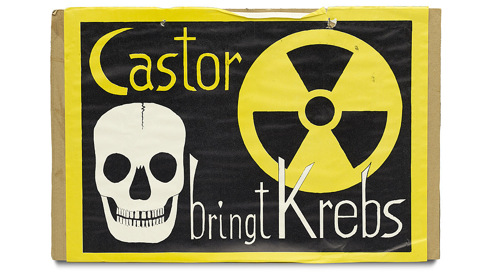 Auf brauner Pappe aufgeklebt: schwarzes Blatt mit gelber Umrandung. Motiv: Totenschädel in schwarz-weiß und das Symbol für Radioaktivität. Dazwischen der Text: 'Castor bringt Krebs'.
