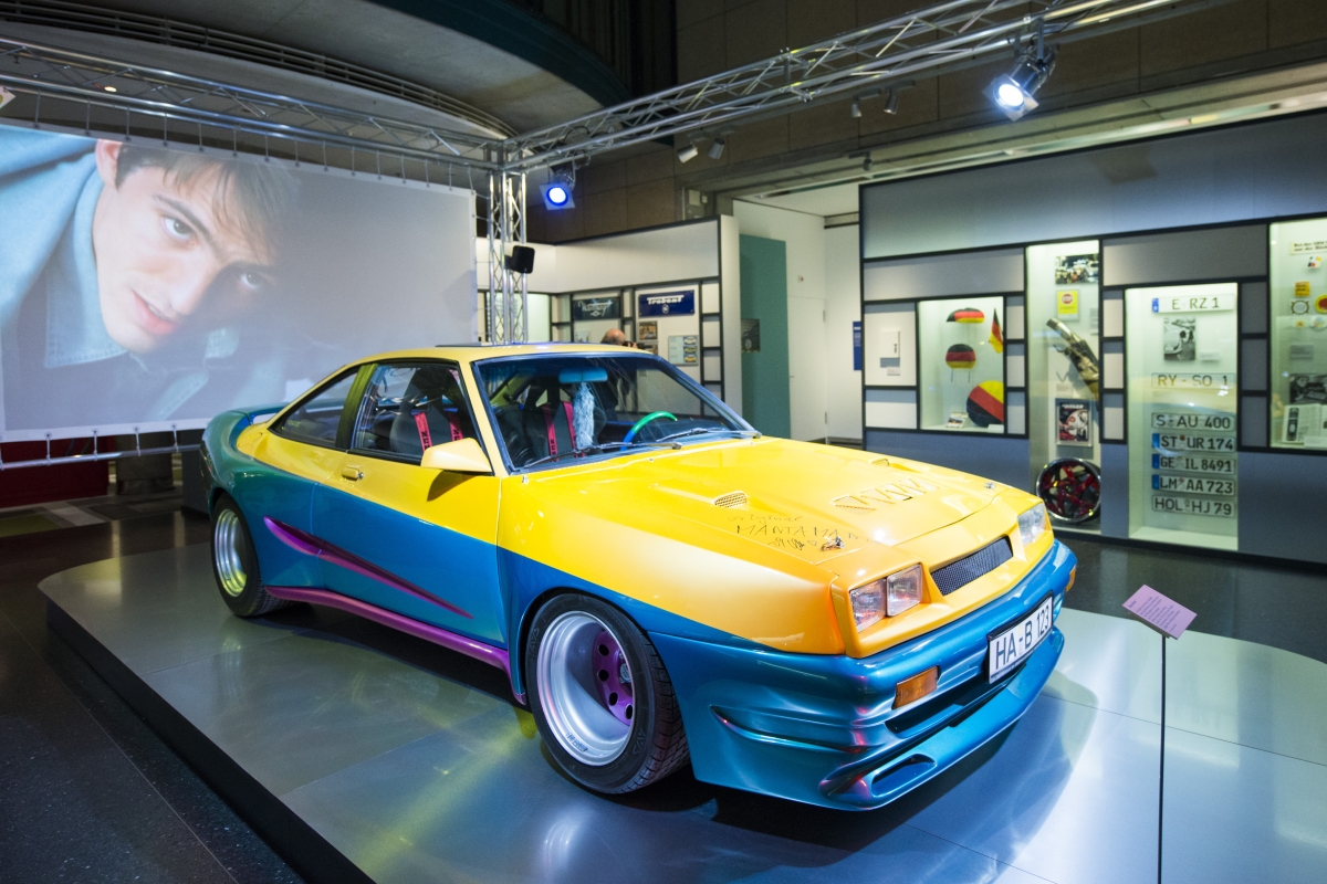 Auf einem flachen Podest in einem Ausstellungsraum steht ein getunter Opel Manta, dahinter eine Großprojektion des Films.