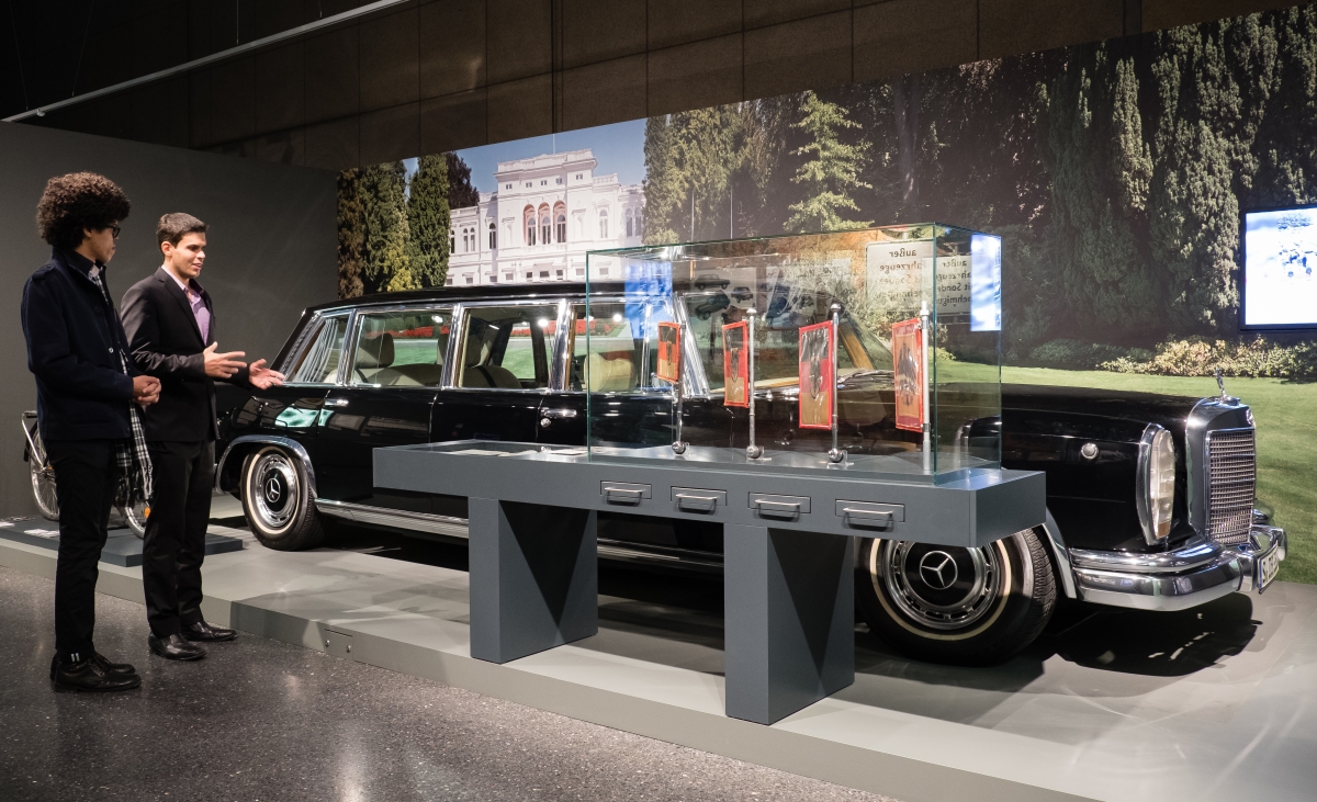 Eine große schwarze Mercedes-Staatslimousine steht in einem Ausstellungsraum vor einem großen Foto der Villa Hammerschmidt, daneben zwei junge Männer.