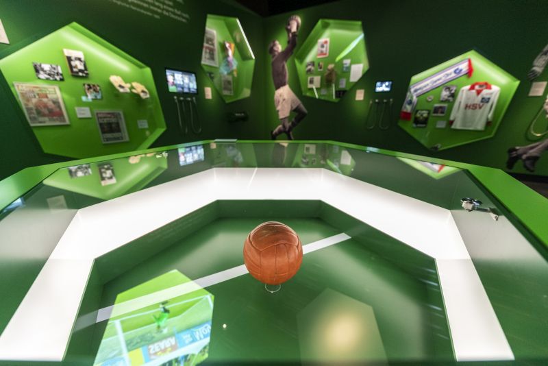 Grün gestrichener Ausstellungsraum zu Rivalität und Respekt mit dem braunen Wembley-Ball in einer Vitrine in der Mitte.