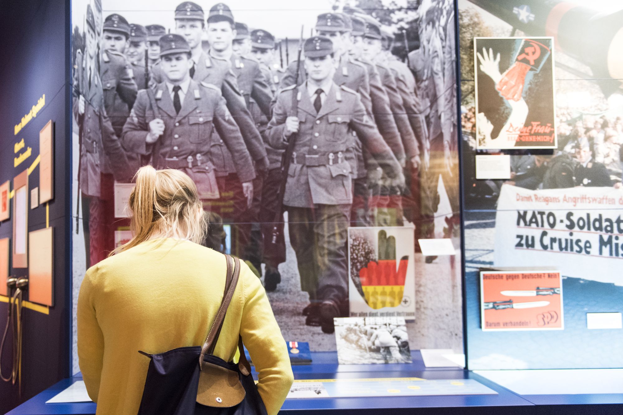 Eine junge Frau in einer gelben Strickjacke mit einer Schultertasche schaut in eine Vitrine, dahinter ein großformatiges Foto von marschierenden Soldaten