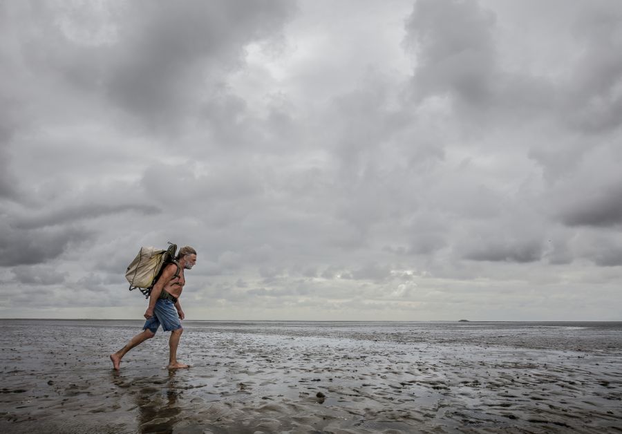 An older man walks through the mudflats with his upper body naked, shorts, and a big backpack.