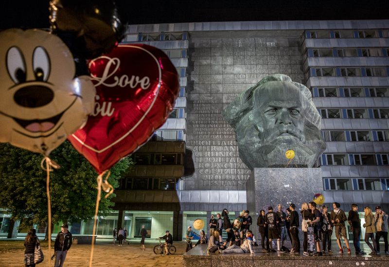 Im Vordergrund sind ein Micky Maus- und ein Herzluftballon zu sehen, im Hintergrund ein Denkmal mit einem großen Marxkopf, auf dessen Sockel Jugendliche feiern. 